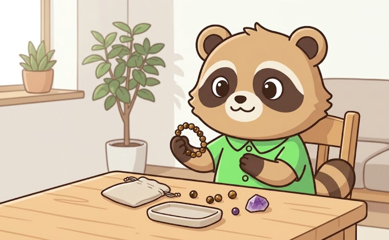 椅子に座った緑のシャツのたぬきが、小粒のタイガーアイブレスレットを手にしながら胸に手を当て、暮らしに馴染むアクセサリーとして天然石を取り入れている様子のイラスト